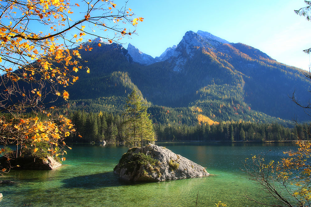 Der Hintersee im Herbst mit Berg Hochkalter im Hintergrund. Vorn eine großer Felsblock im Wasser, auf dem ein Nadelbaum wächst.