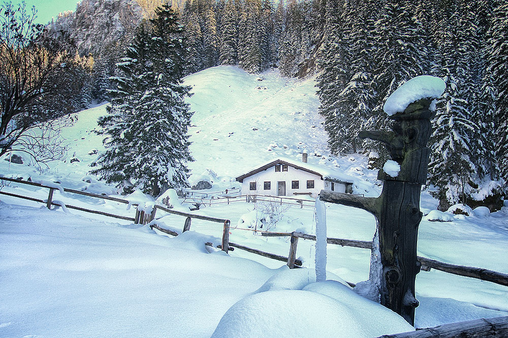 Eine Hütte steht an einem verschneiten Berg mit Fichten, vorn ein Holzzaun, rechts eine alte Wasserpumpe aus Holz
